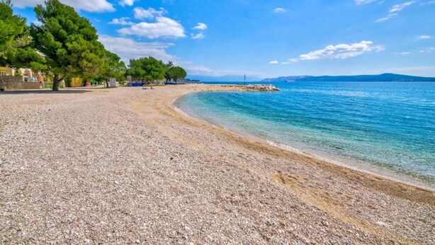 Platja de petits còdols amb aigua blava i cristal·lina, arbres i una vista panoràmica del mar.