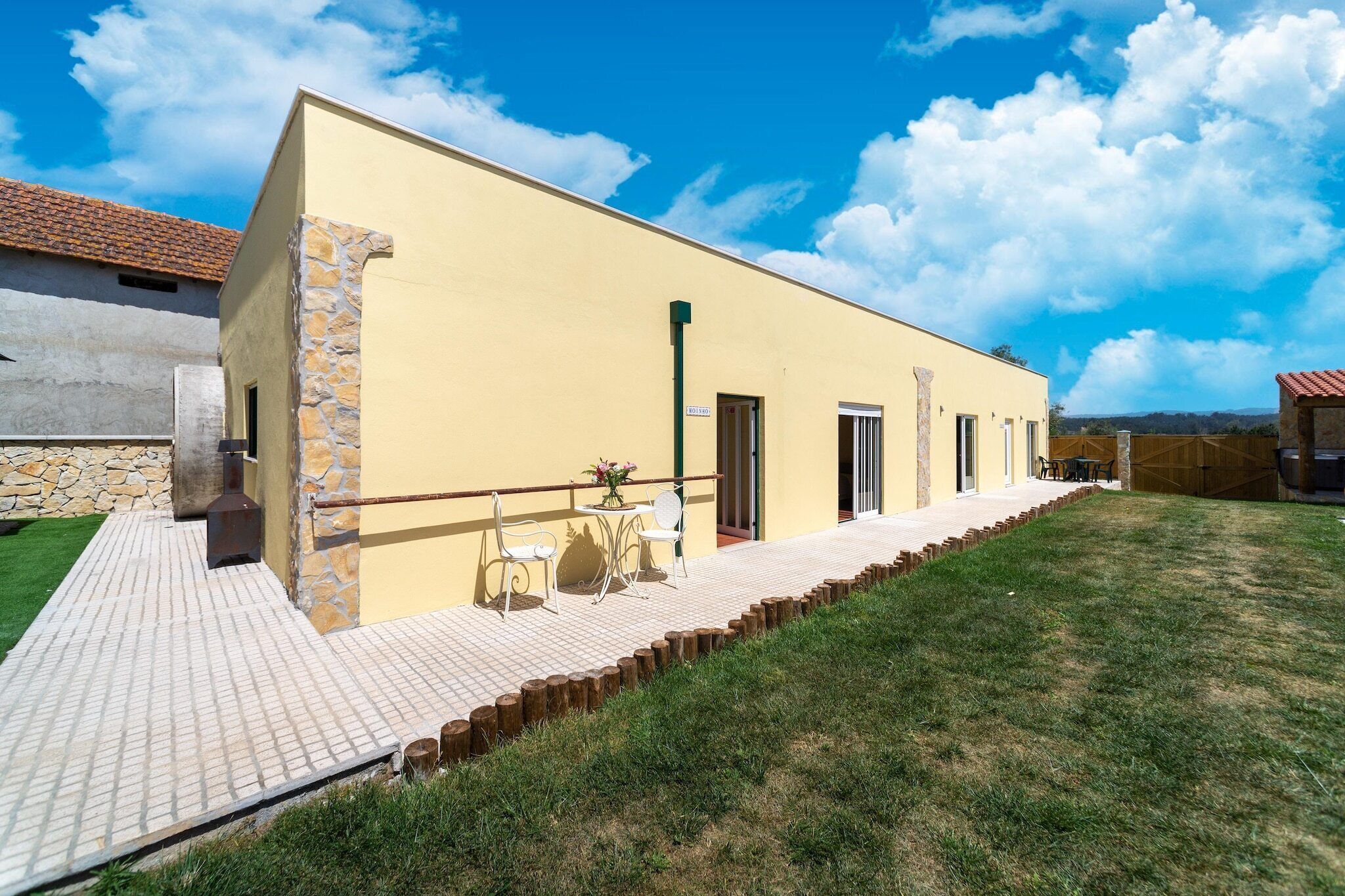 Um edifício amarelo com fachada de pedra, um pátio pavimentado com mobiliário de exterior e uma área ajardinada.