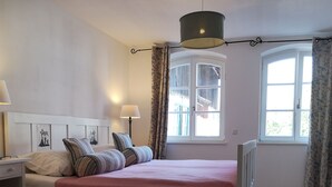 Ett sovrum med en säng, sängbord, en lampa, ett fönster med gardiner, ett element och en mönstrad matta.