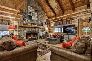 A cozy log cabin interior with a stone fireplace, leather furniture, and a mounted television.