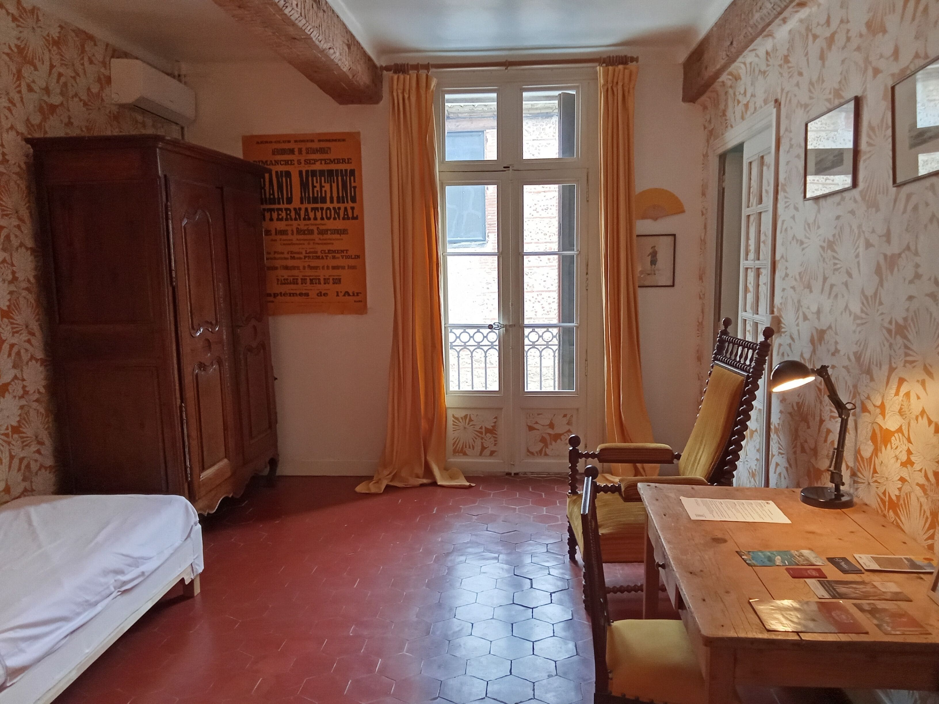 A room with a bed, a wooden chair, a desk with papers, a lamp, and a window with curtains.