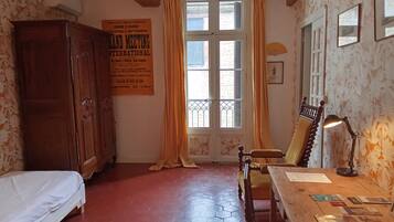 A room with a bed, a wooden chair, a desk with papers, a lamp, and a window with curtains.