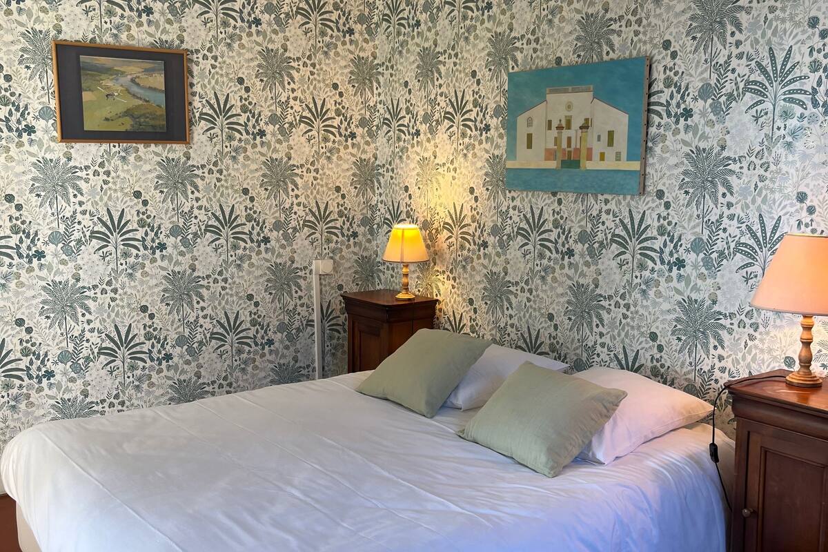 A neatly made bed with white bedding, a wooden nightstand with a lamp, and a wall with floral wallpaper.