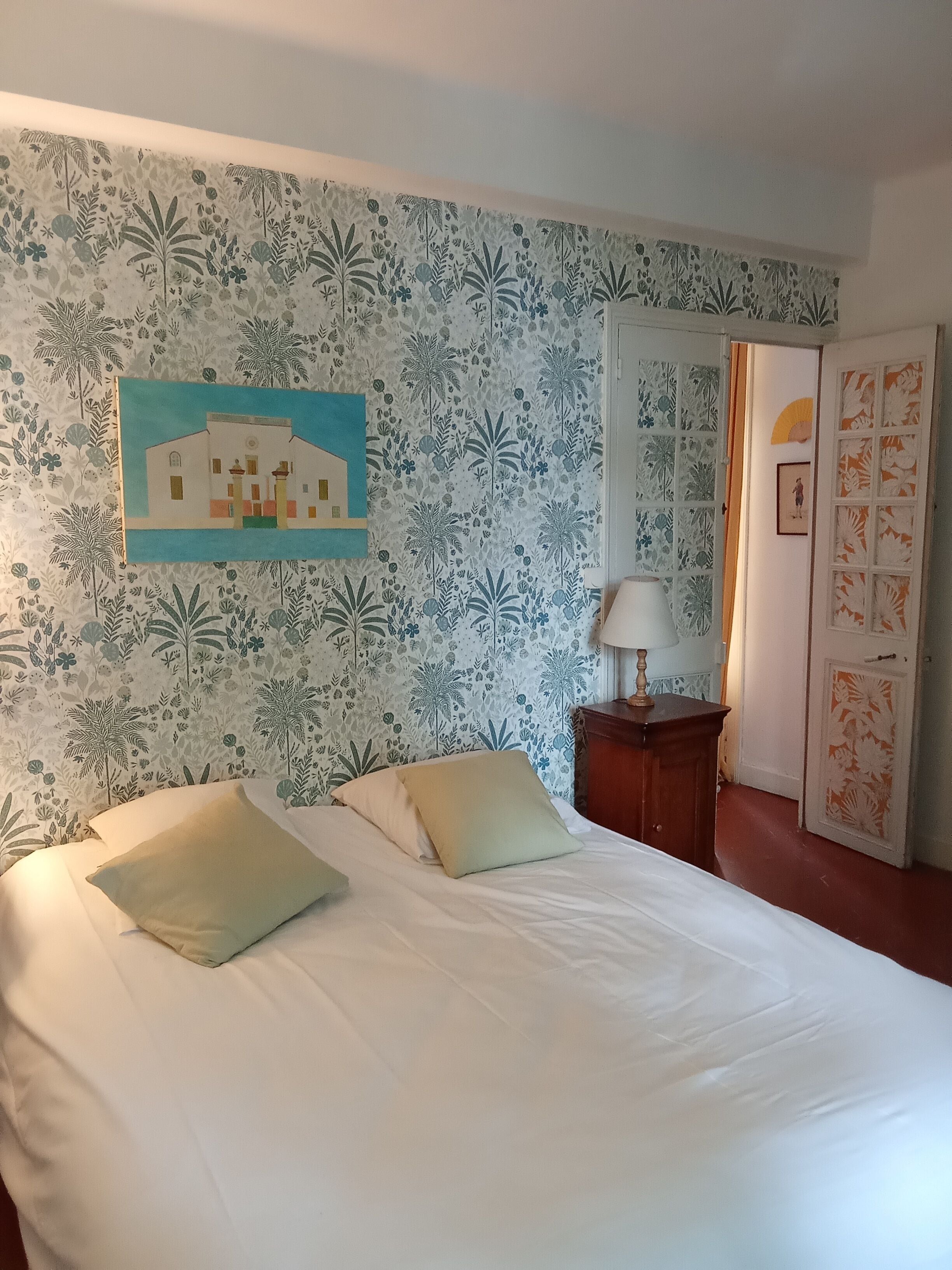A bedroom with a bed, pillows, a nightstand, a lamp, a door with a floral pattern, and a wall with a floral wallpaper and a painting.