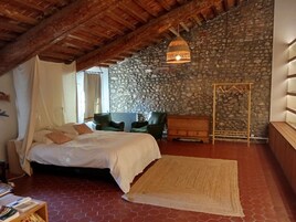 A bedroom with a stone wall, a bed with white bedding, a green armchair, and a hanging light fixture.
