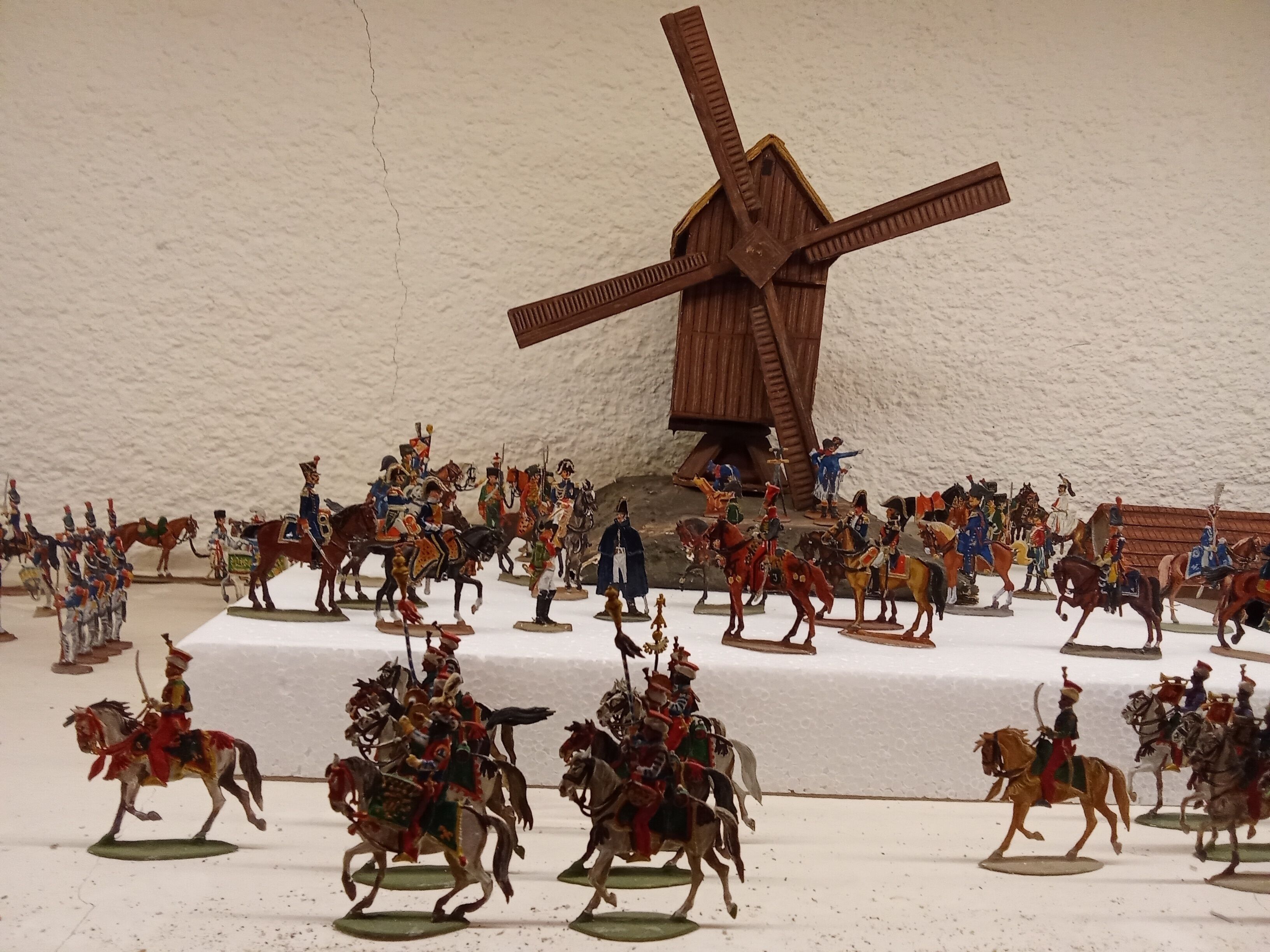 Une maquette représentant une scène de bataille historique, avec des figurines miniatures à cheval, un moulin à vent en bois en arrière-plan et un mur blanc à la texture particulière.