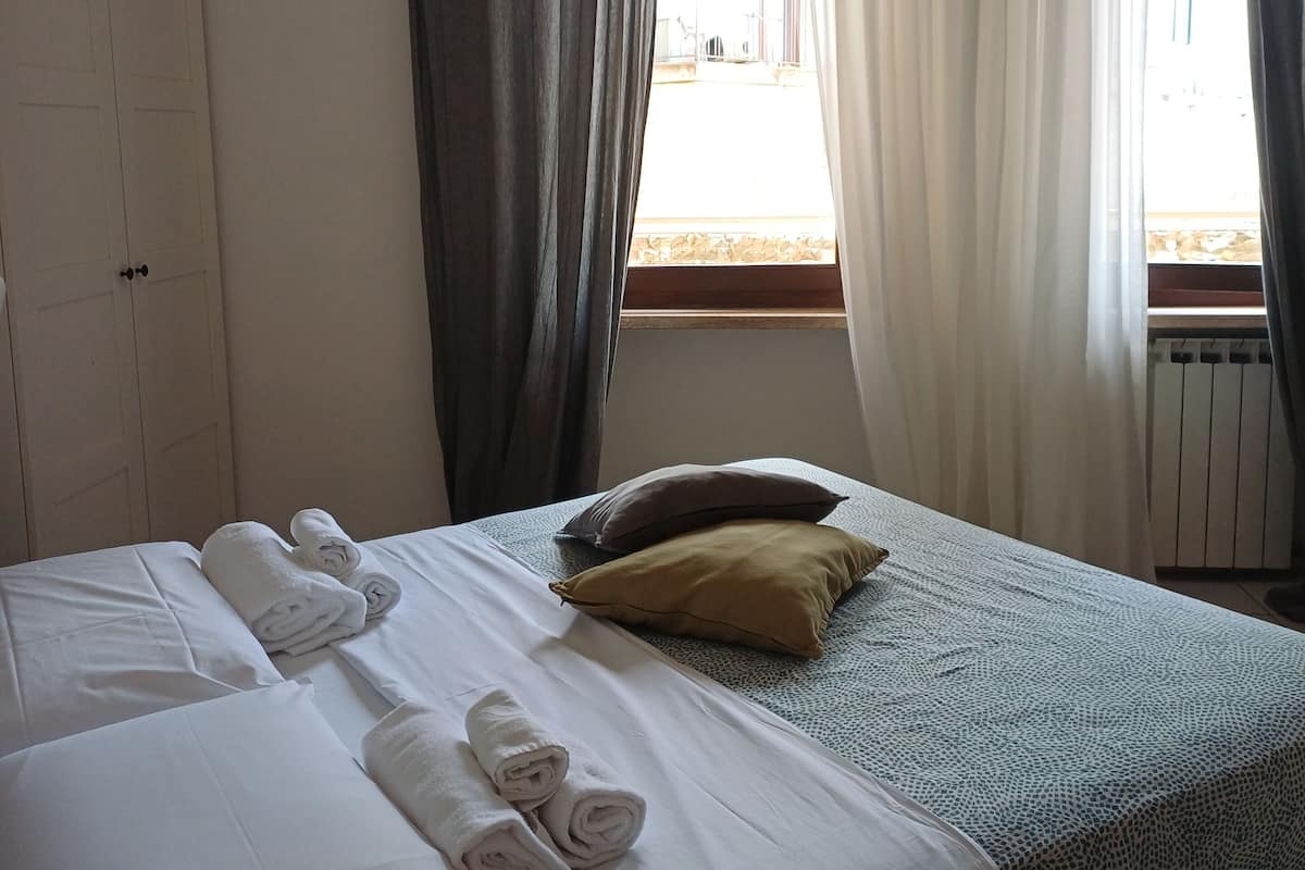 Una cama bien hecha con toallas enrolladas, una ventana con cortinas y un radiador.