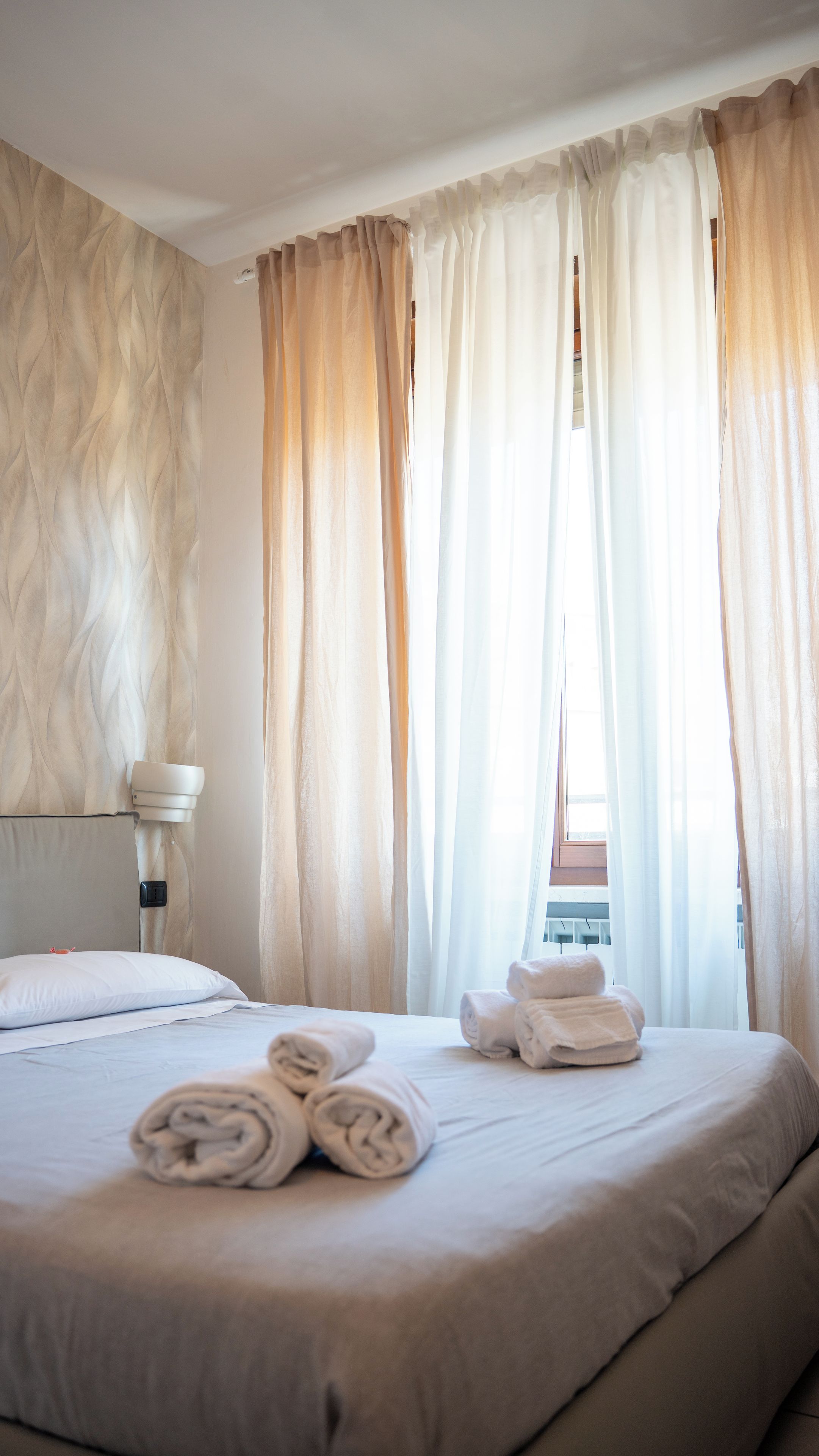 A hotel room with a bed, rolled towels, a bedside lamp, and sheer curtains.