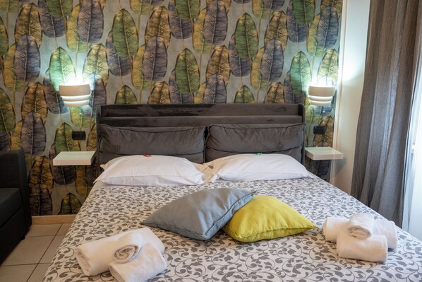 Una habitación de hotel con cama, almohadas, mesita de noche, lámpara y ventana con cortinas.