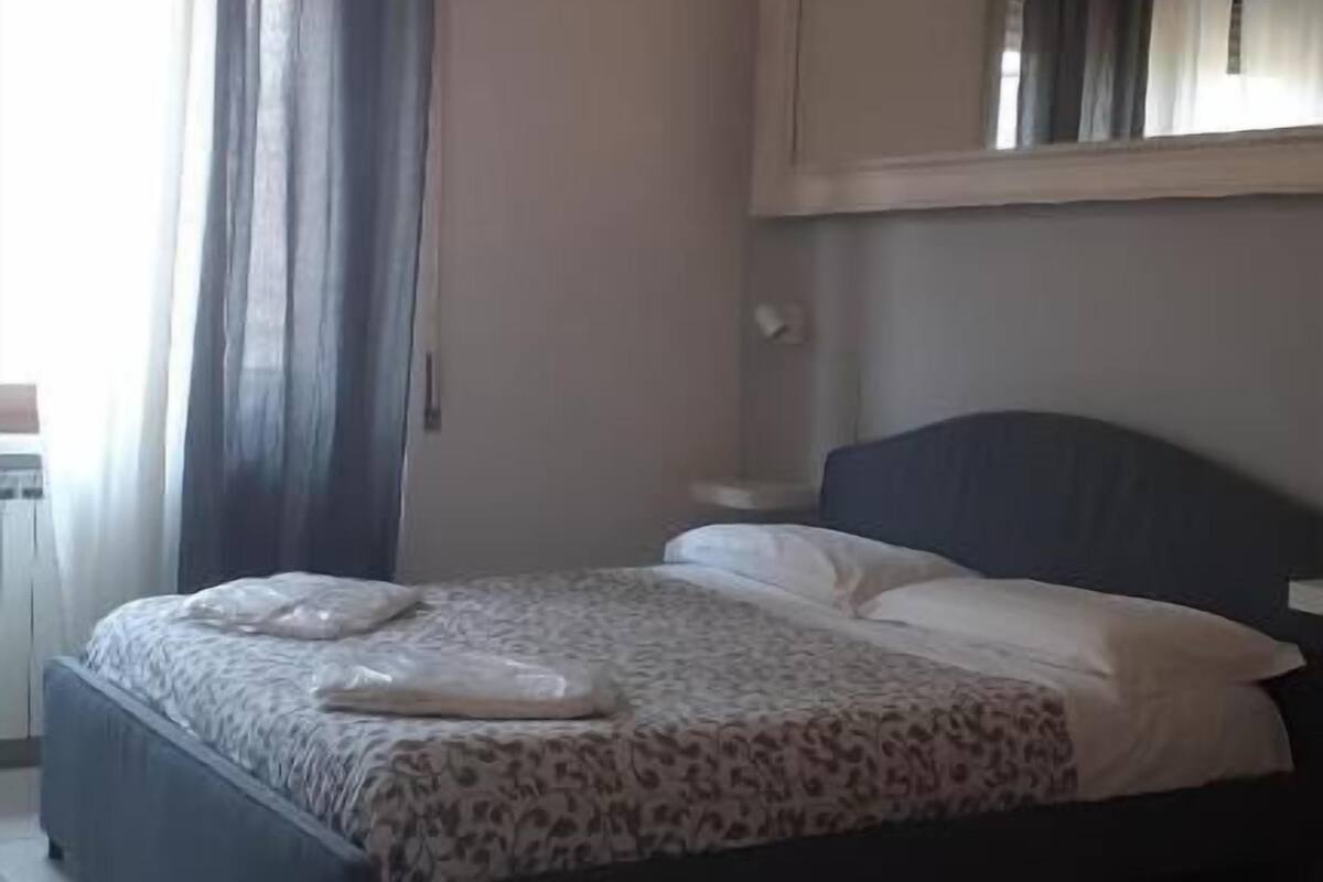 Un dormitorio con cama, lámpara colgante, ventana con cortinas y espejo en la pared.