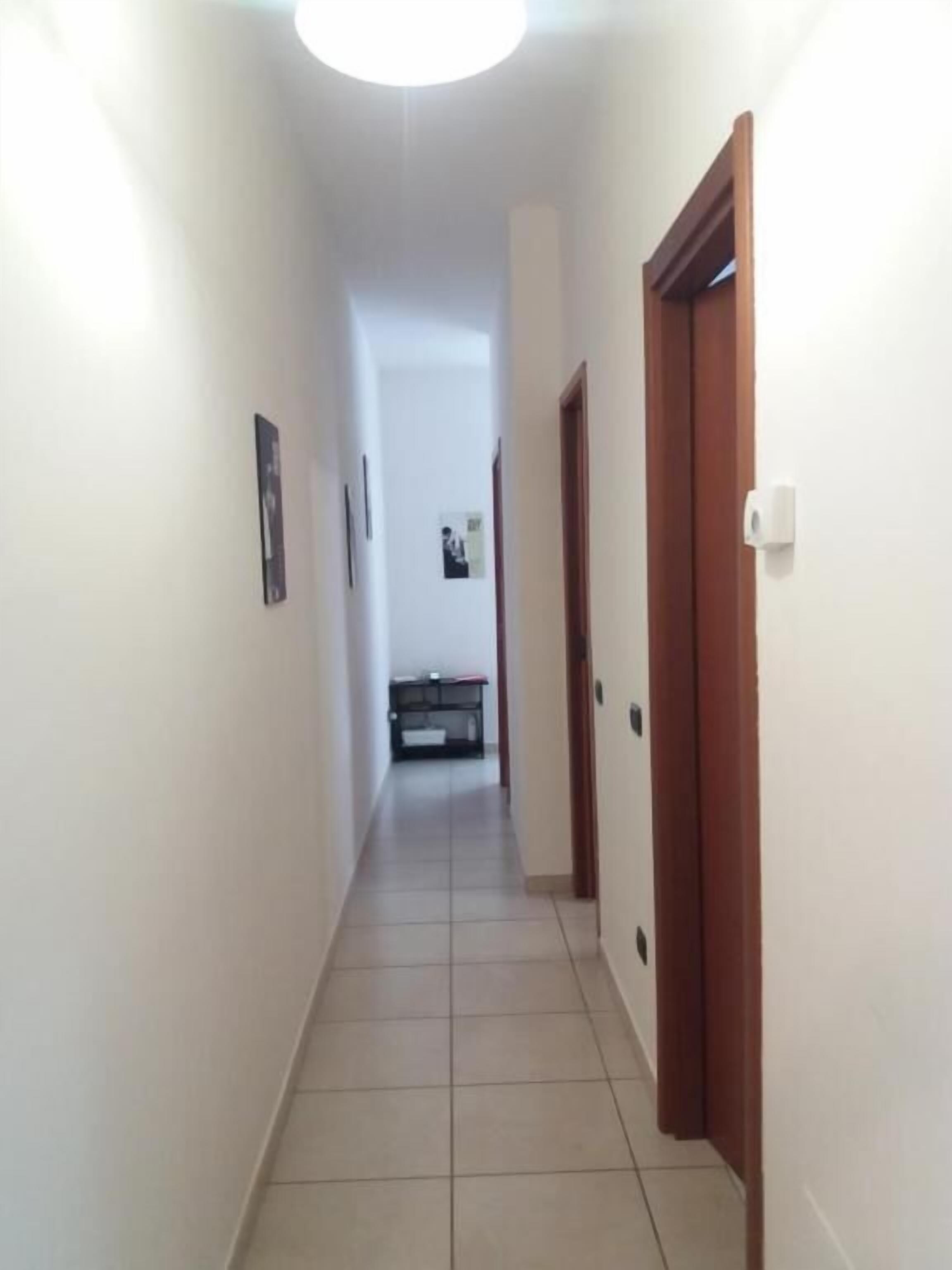 Un pasillo estrecho con paredes blancas y suelo de baldosas, que conduce a una habitación con un escritorio y una silla.