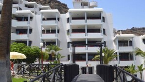 A multi-story hotel with balconies, a central entrance, and a view of a rocky hillside. - El Cardenal (Mogan)