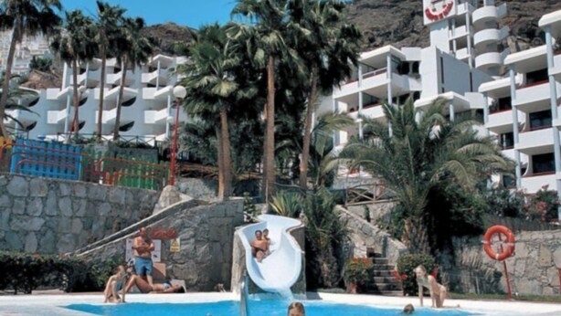 A swimming pool area with a water slide, palm trees, and a multi-story building in the background. - El Cardenal (Mogan)