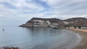 A coastal area with a sandy beach, calm sea, and a large building complex with terraces. - El Cardenal (Mogan)