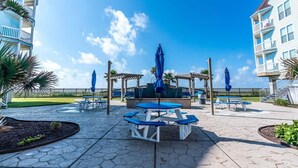 A patio area with picnic tables and umbrellas, surrounded by buildings with balconies. - Easy Beach Access! Charming Beachside Condo! (Galveston)