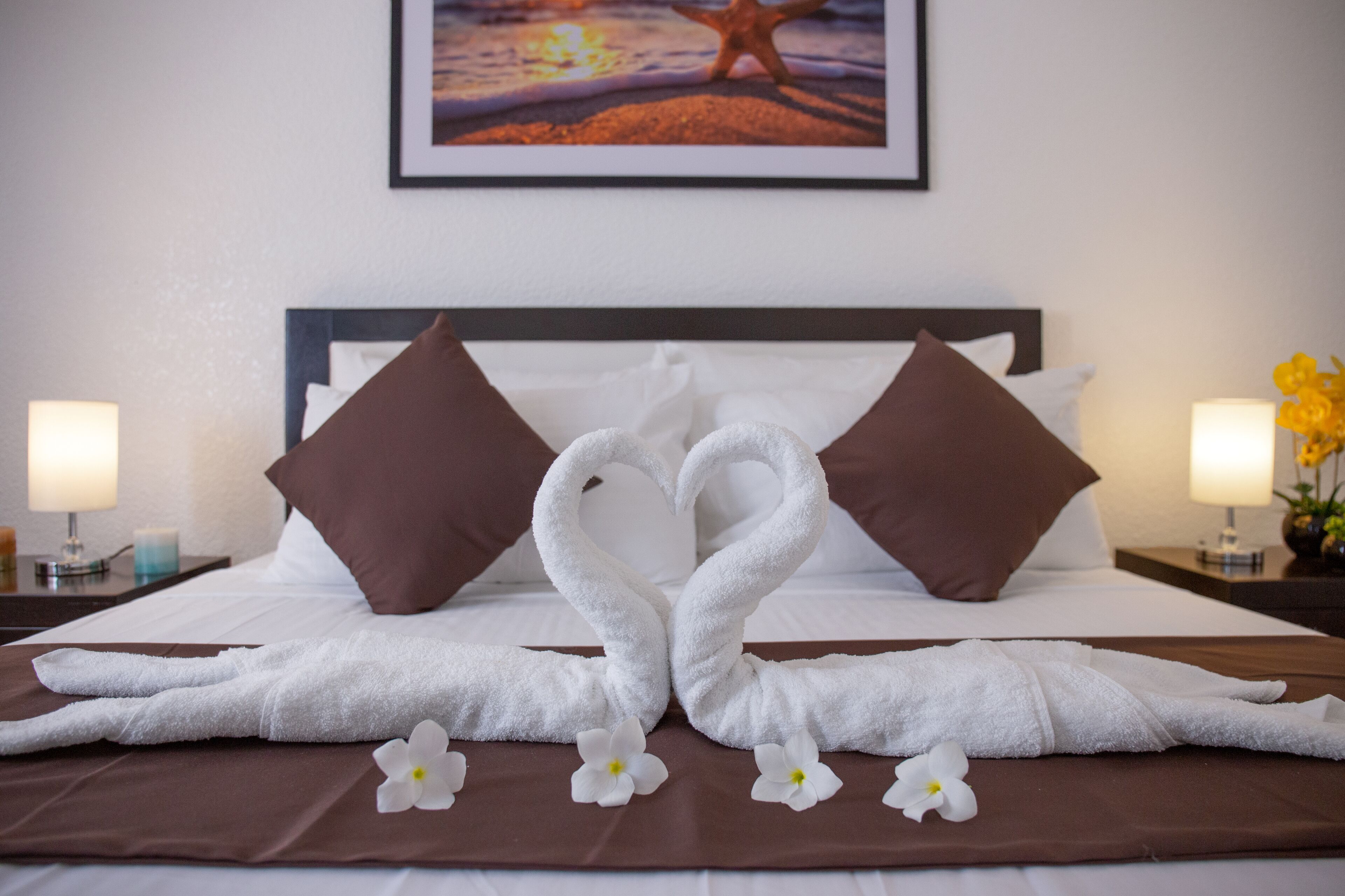 A neatly made bed with two towels arranged in the shape of swans, white flowers, and brown pillows.