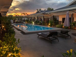 A rectangular pool with a metal ladder, surrounded by lounge chairs and umbrellas, set against a backdrop of a building with a tiled roof and a sunset sky. - Aila Homestay (Panglao)
