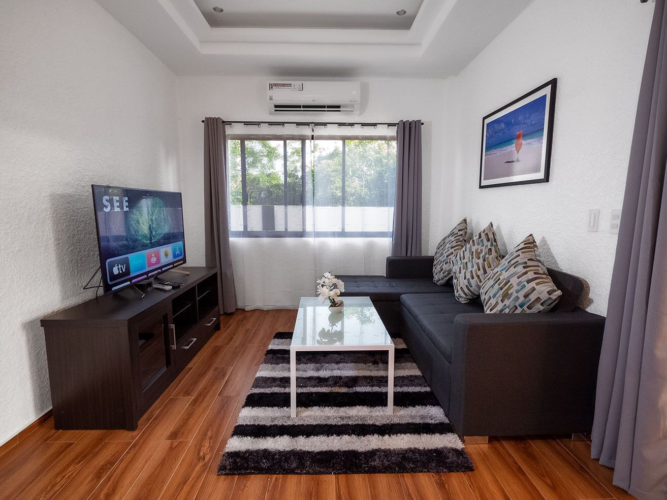 A modern living room with a flat-screen TV, a glass coffee table, a dark sofa with patterned cushions, and a large window with curtains.