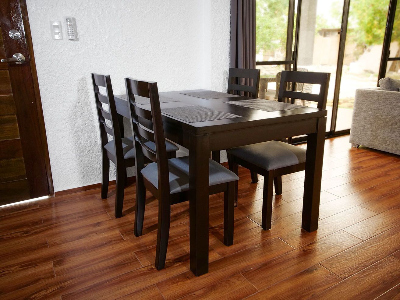 A dining area with a wooden table and chairs, a white textured wall, and a view through large windows.