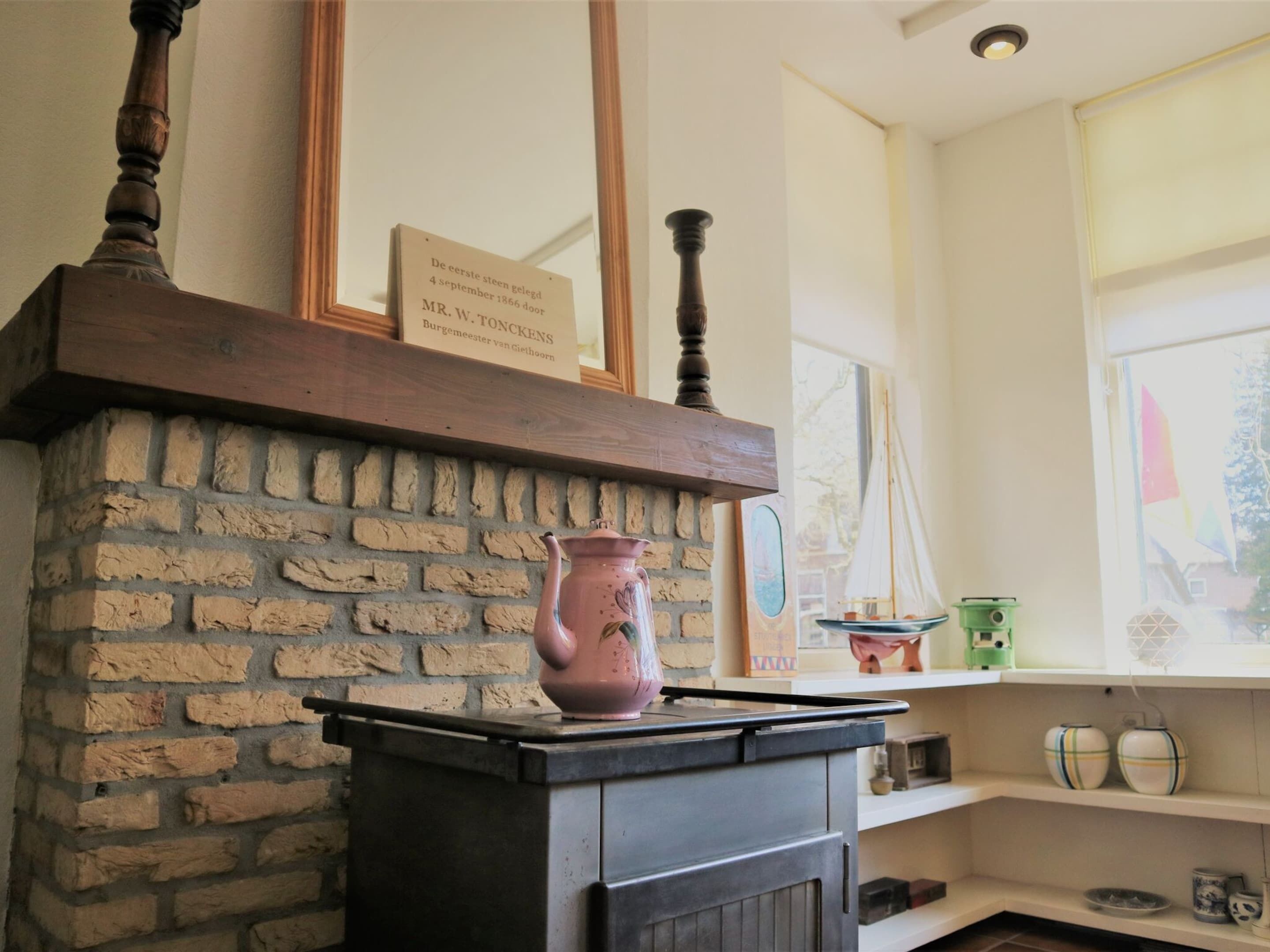 A kitchen with a brick wall, a wooden mantel, a pink vase, and a window with a view of a sailboat.