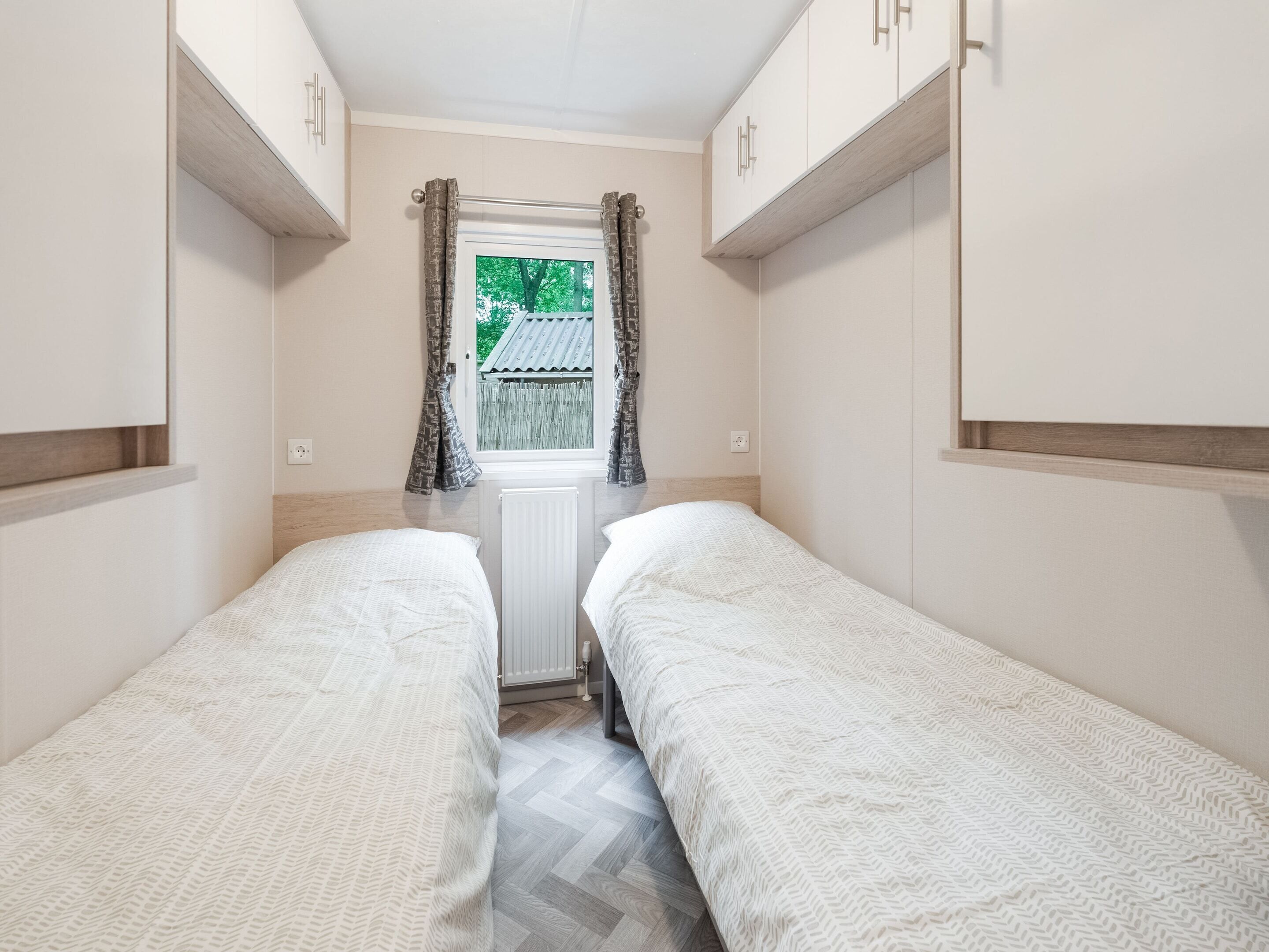 Una habitación pequeña y estrecha con dos camas individuales, una ventana con cortinas y un radiador.
