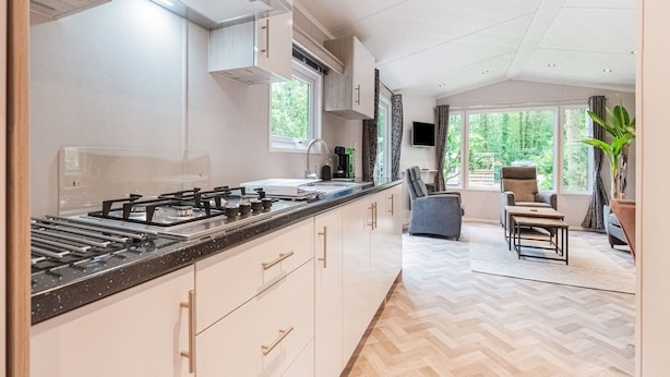Et moderne køkken med en lang bordplade, indbyggede skabe og en gasblus. Der er et spiseområde med et bord og stole, samt et stort vindue der giver udsigt til grønne omgivelser.