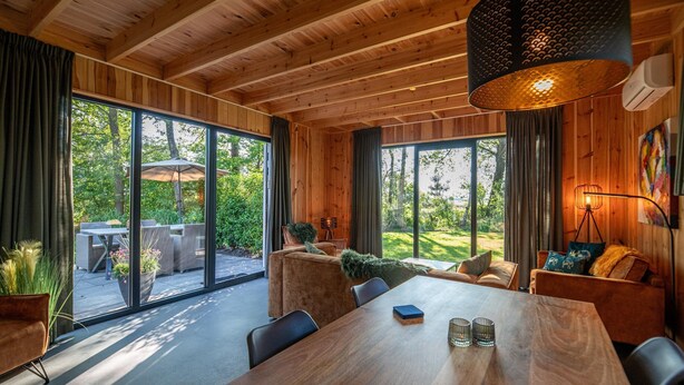 A modern interior with a large dining table, wooden ceiling, and large windows offering a view of the outdoors. - Pleasant Nature Lodge in Dalerveen (Dalerveen)
