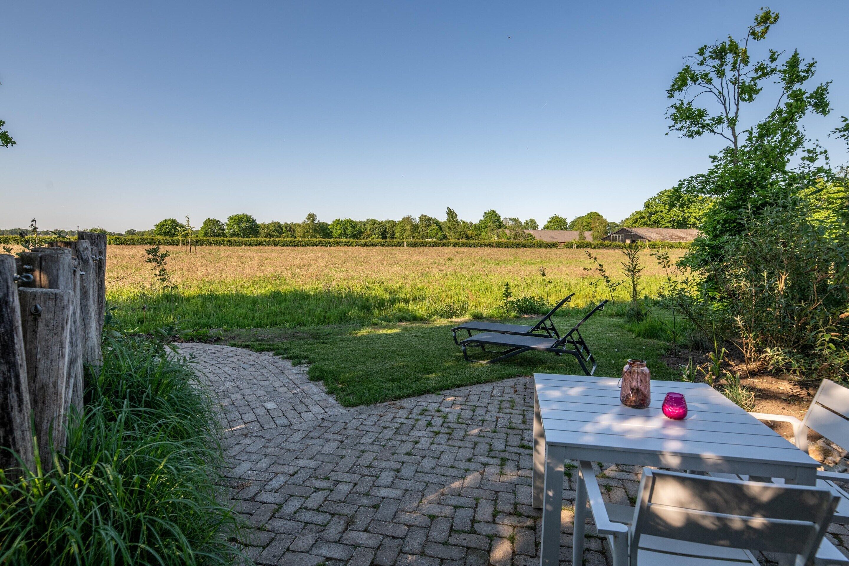 A paved patio with a table and chairs, a wooden fence, and a view of a field and distant buildings.