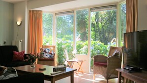 A living room with a sofa, a table with a vase of flowers, a TV, and a view of a garden through large windows. - Holiday Home Buitenplaats Berg With 5 Rooms (Lemele)