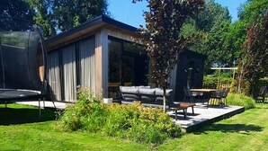 A modern outdoor seating area with a glass enclosure, a wooden deck, and a small tree. - Peaceful Chalet Close to Norg (Haulerwijk)