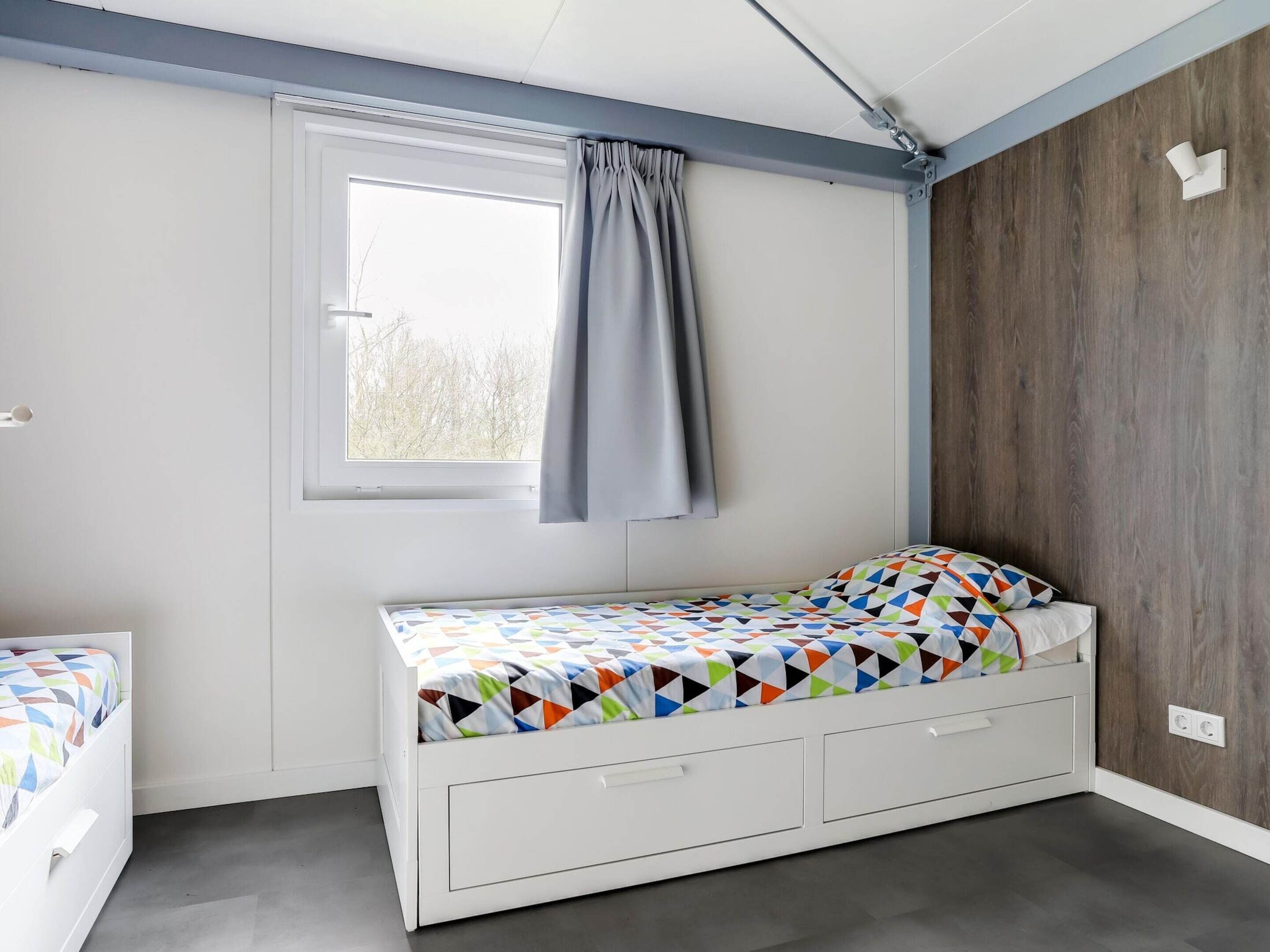 A bedroom with a single bed, a window with curtains, a wooden wall, and a white ceiling.