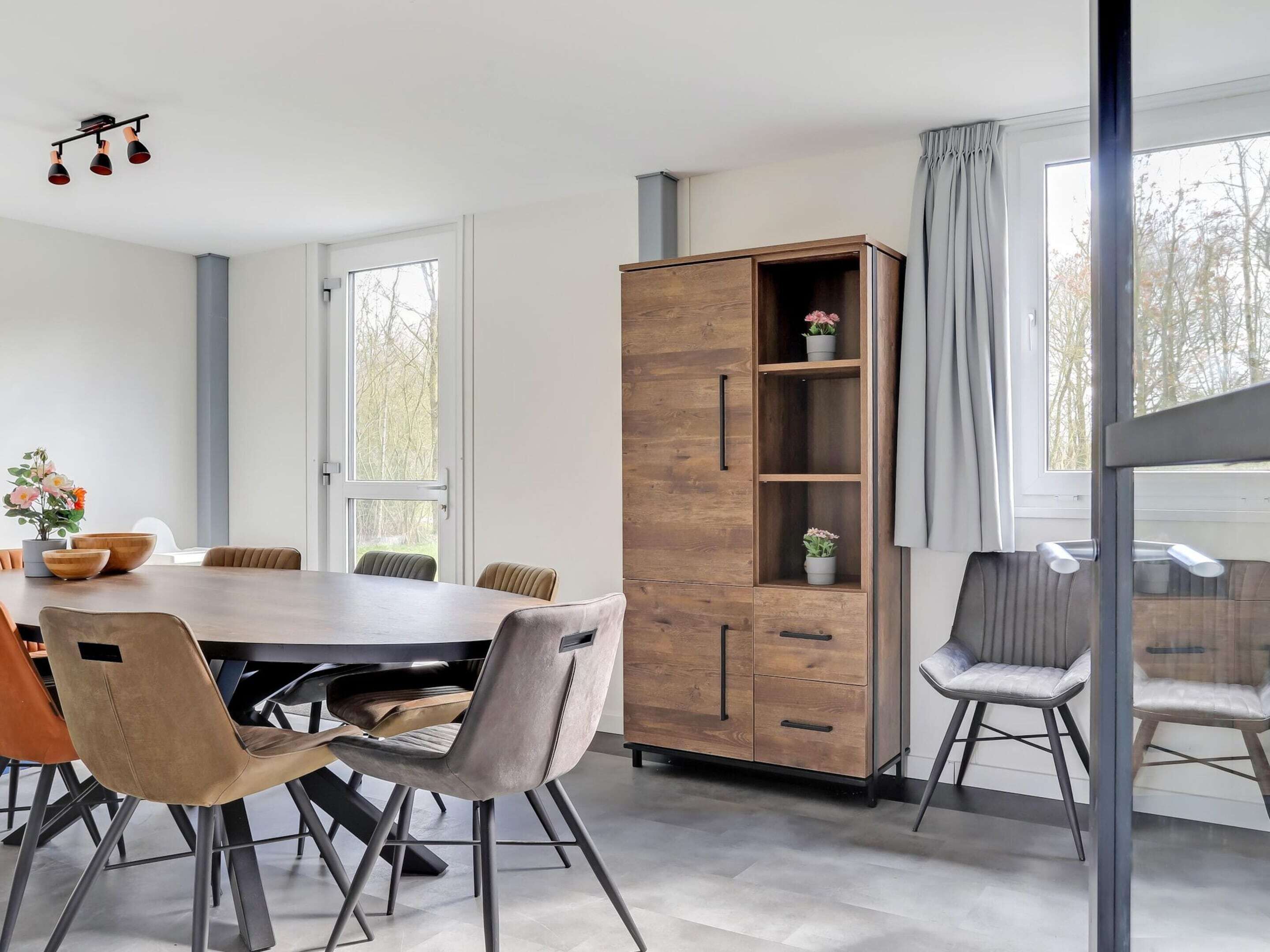 A modern dining area with a wooden table and chairs, a tall cabinet with shelves, and large windows with curtains.