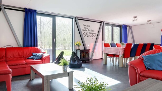 A modern living room with a red sofa, a wooden coffee table, and a dining area with red and blue chairs. - Spacious Holiday Home in Noordwolde With Garden (Noordwolde)
