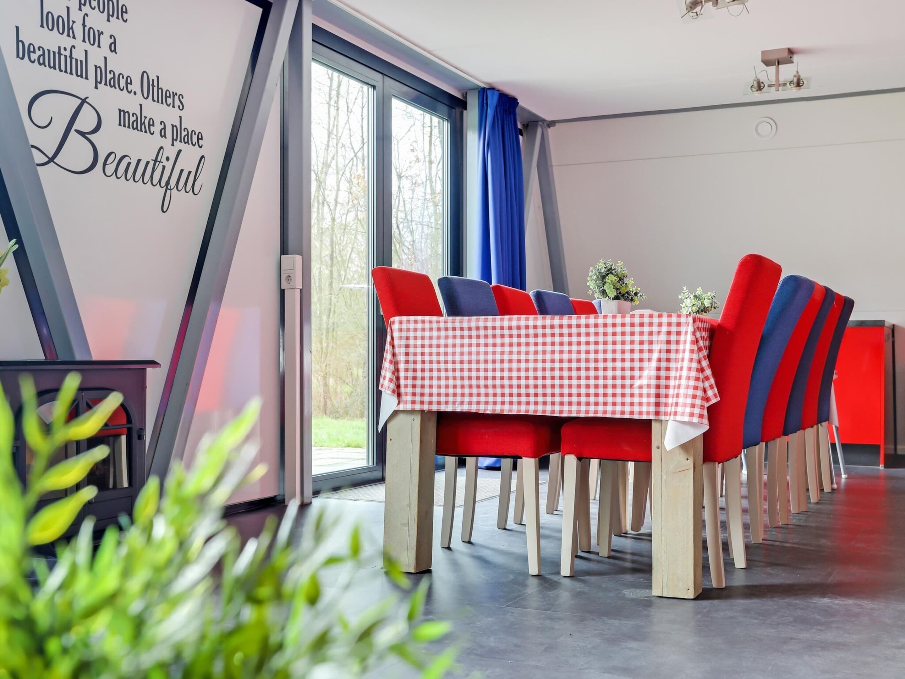 A dining area with a checkered tablecloth, red and blue chairs, and a view of trees through large windows.