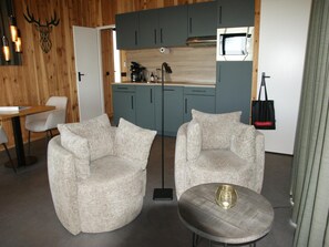 Une cuisine moderne avec des meubles de couleur turquoise, un four à micro-ondes et une machine à café. On y trouve deux fauteuils beiges et une petite table ronde agrémentée d’un objet décoratif.