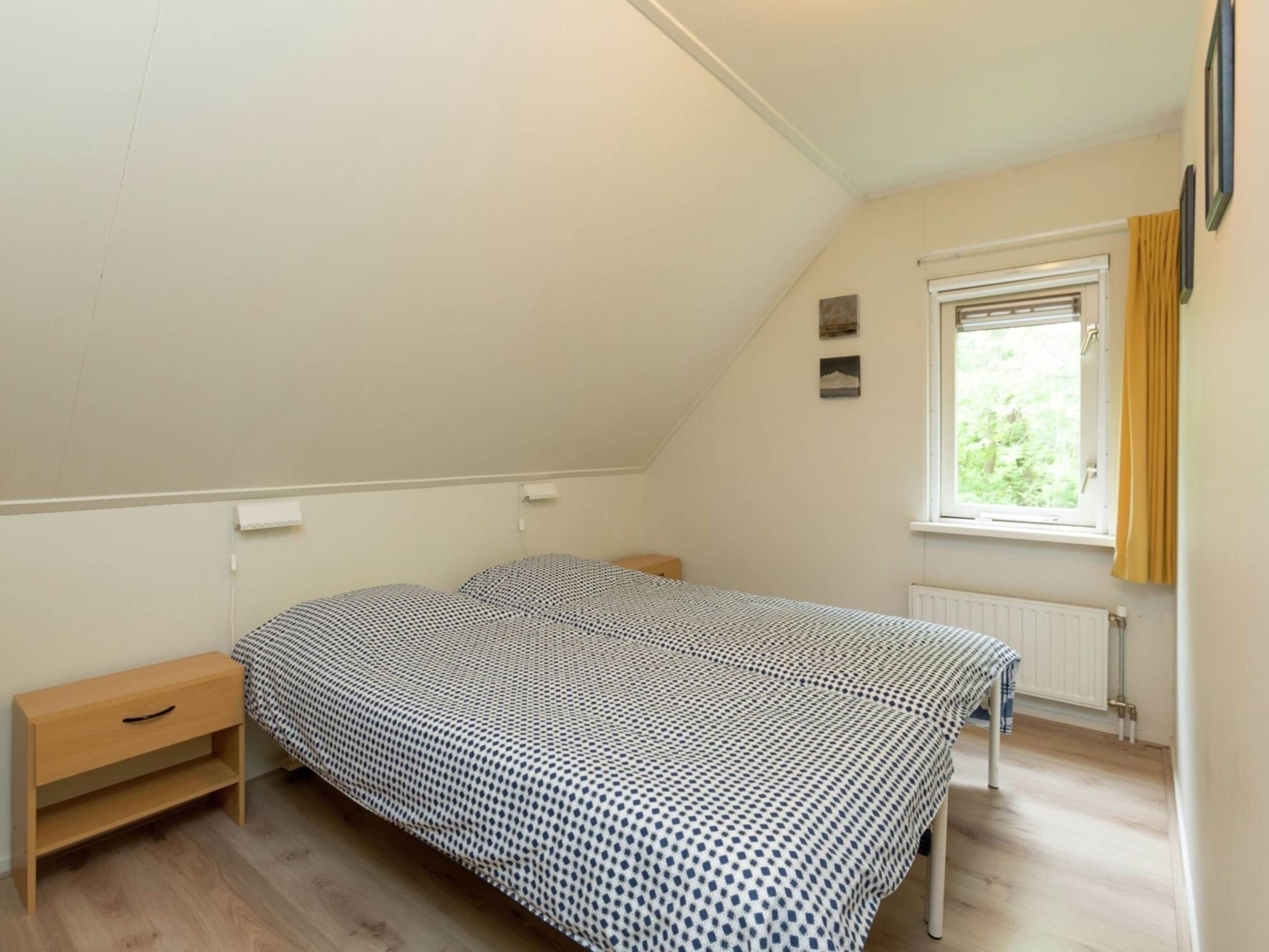 A small, single-bed room with a sloped ceiling, a window with curtains, a radiator, and a small bedside table.