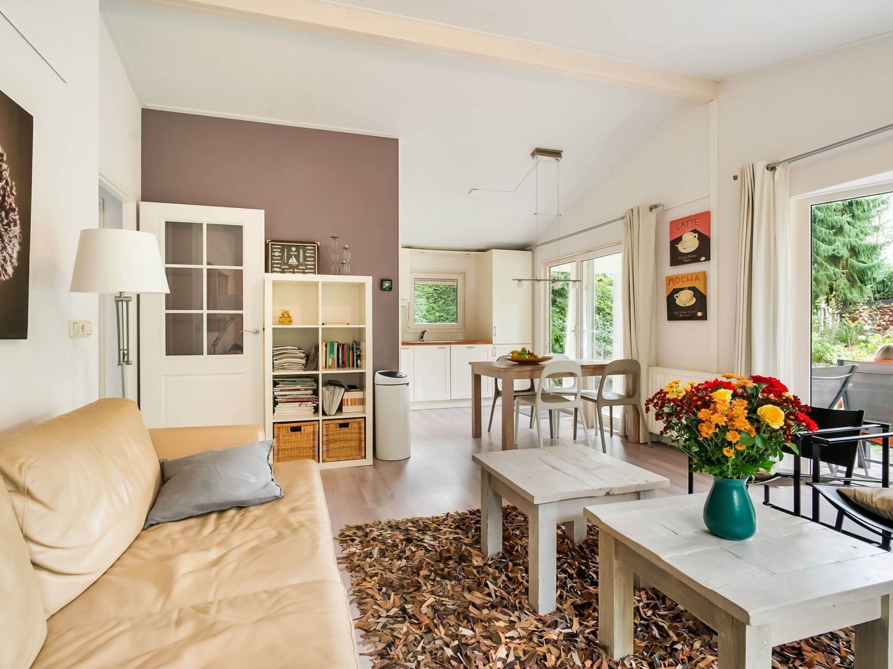 A modern living room with a beige sofa, a coffee table, and a dining area with a table and chairs. There is a bookshelf filled with books and decorative items, and a window with a view of greenery outside.