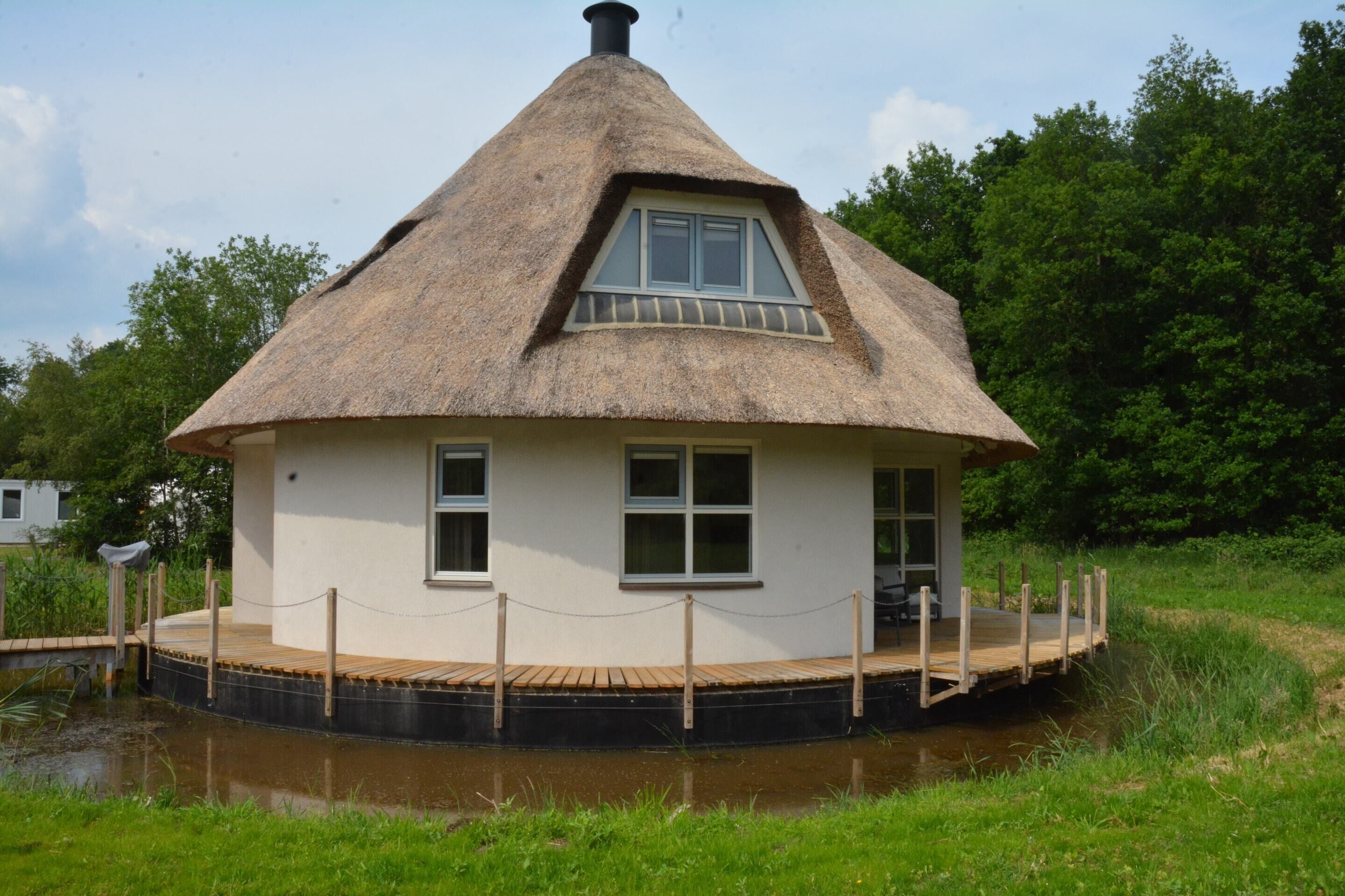 Una casa con techo de paja y terraza de madera, ubicada junto a una masa de agua, rodeada de follaje.