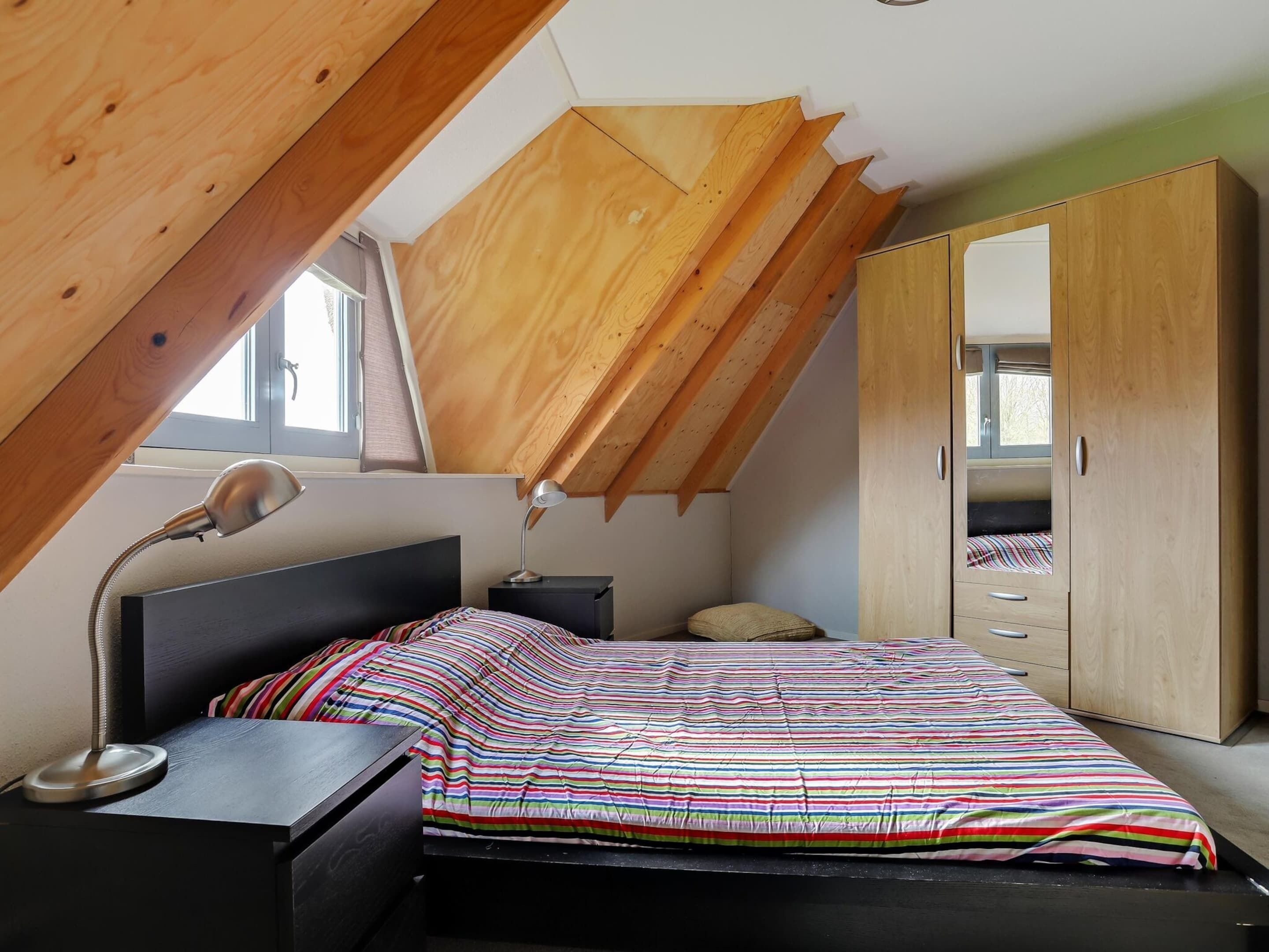 A bedroom with a sloped ceiling, a bed with a striped bedspread, a nightstand with a lamp, and a wardrobe.