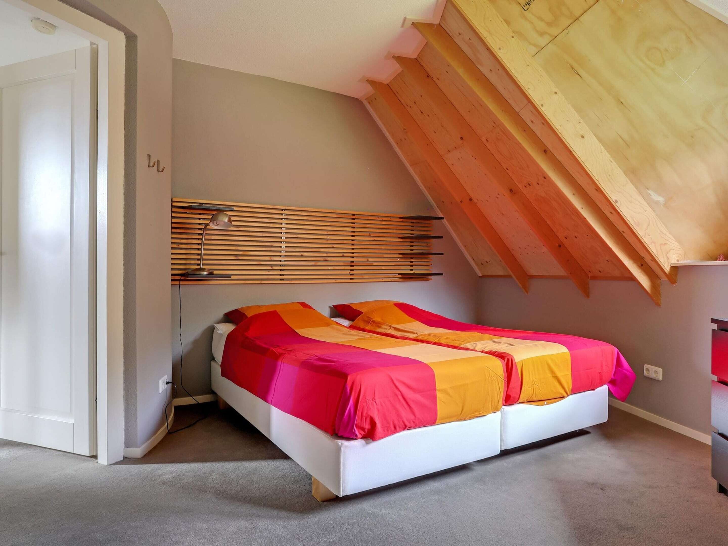 Un dormitorio con techo inclinado, una cama con colcha colorida, cabecera de madera y una lámpara fijada a la pared.