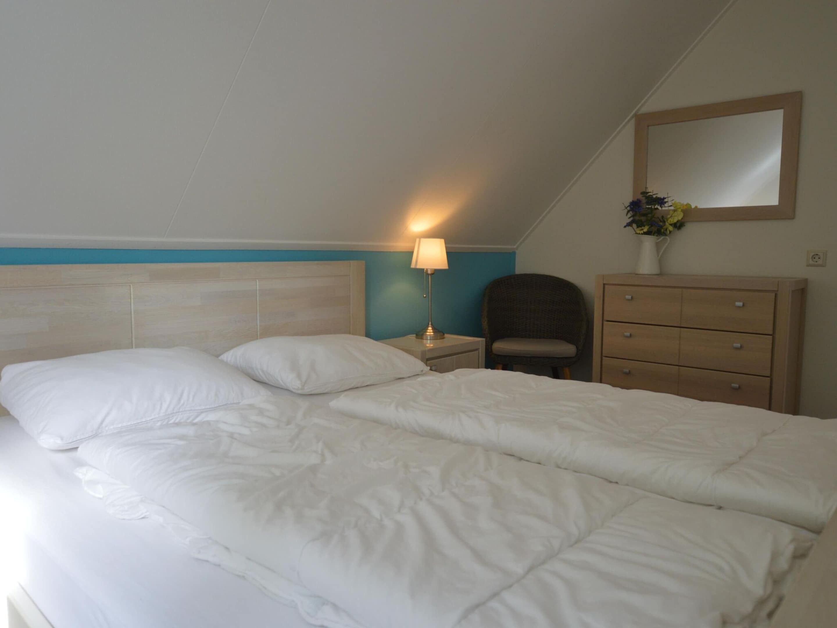 A neatly made bed with white linens, a bedside lamp, a chair, and a dresser in a hotel room.