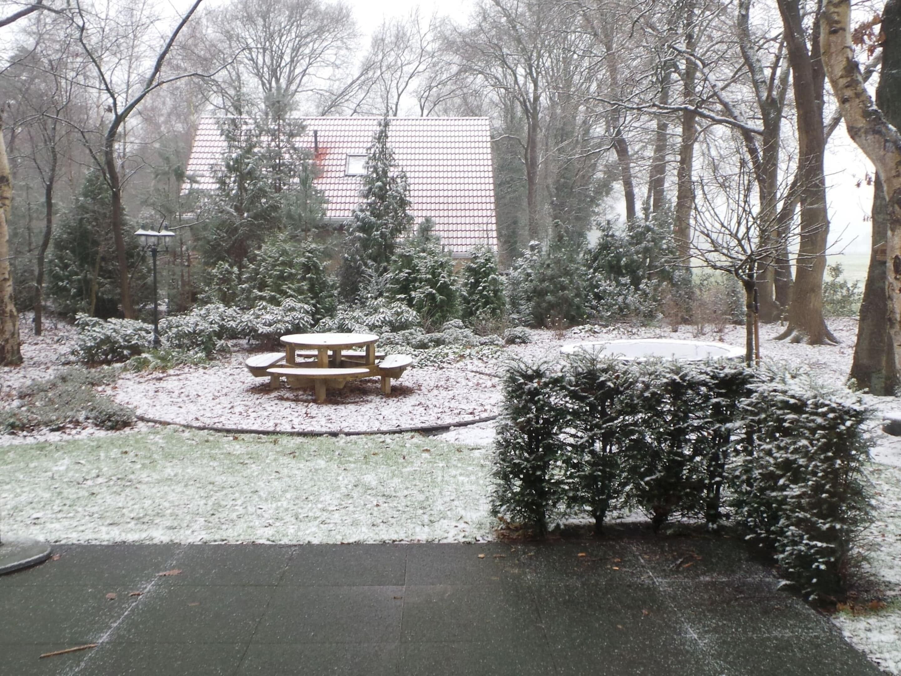 Ein mit Schnee bedeckter Garten mit einem Picknicktisch und Bänken, umgeben von Bäumen und einem Gebäude im Hintergrund.