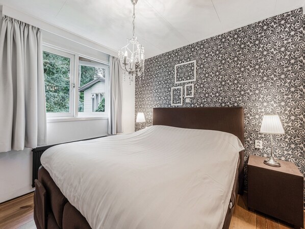 A bedroom with a large bed, a chandelier, a nightstand, and a window with curtains.