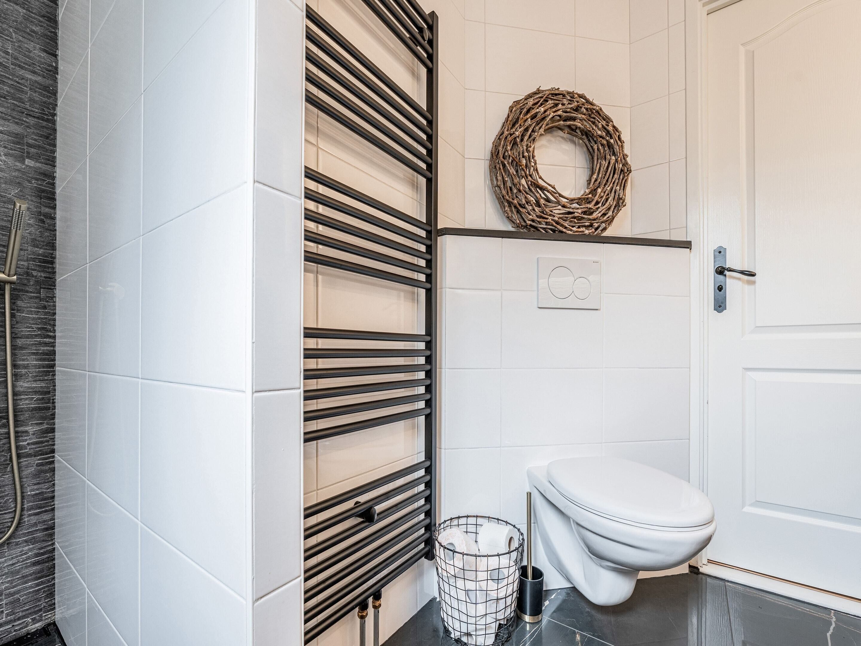 A bathroom with a white toilet, a shower with a handheld showerhead, a towel rack with a woven wreath, and a waste basket.