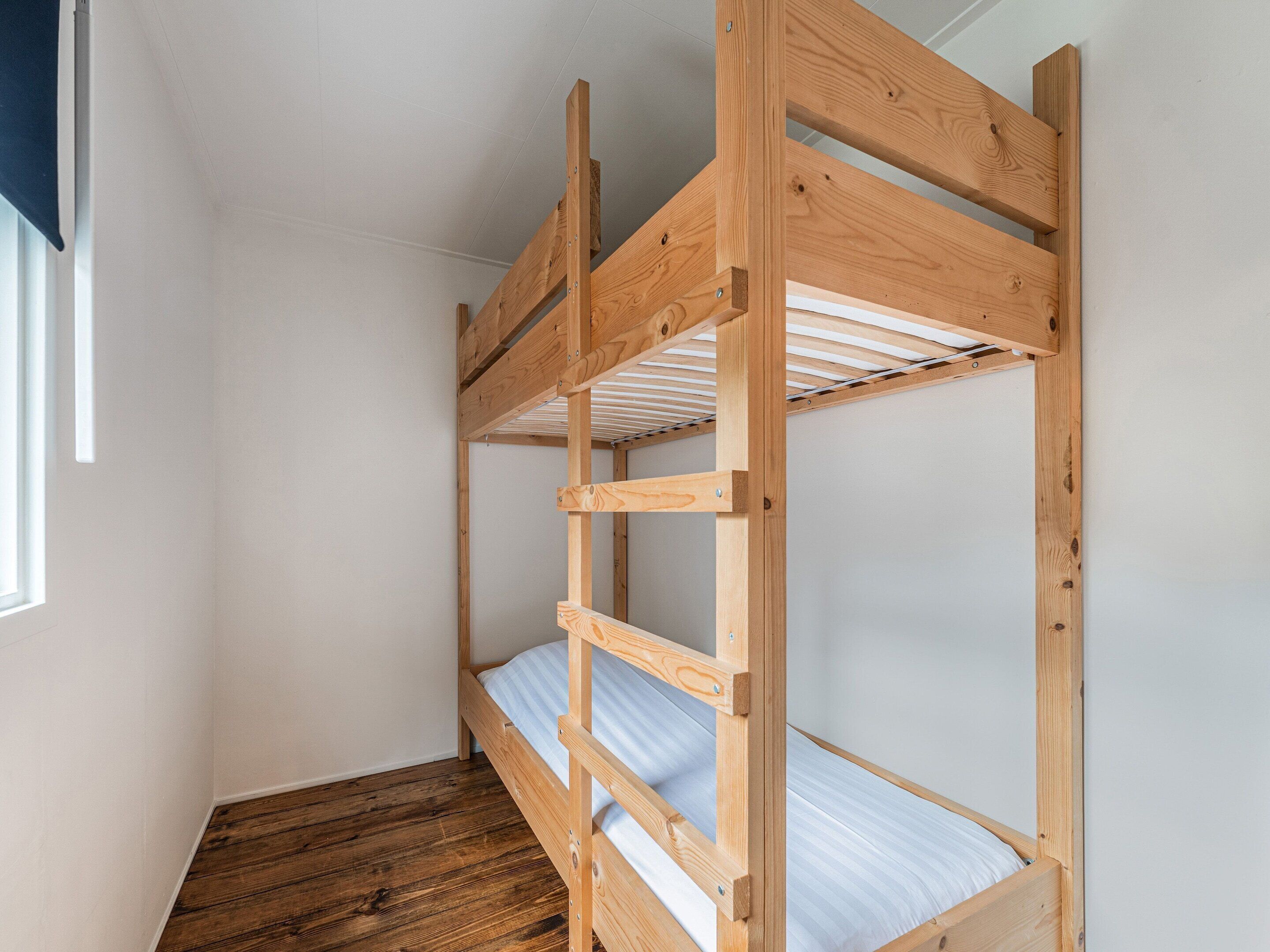A bunk bed with a wooden frame and a metal ladder, built into a corner of a room with white walls and a wooden floor.