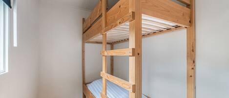 A bunk bed with a wooden frame and a metal ladder, built into a corner of a room with white walls and a wooden floor.