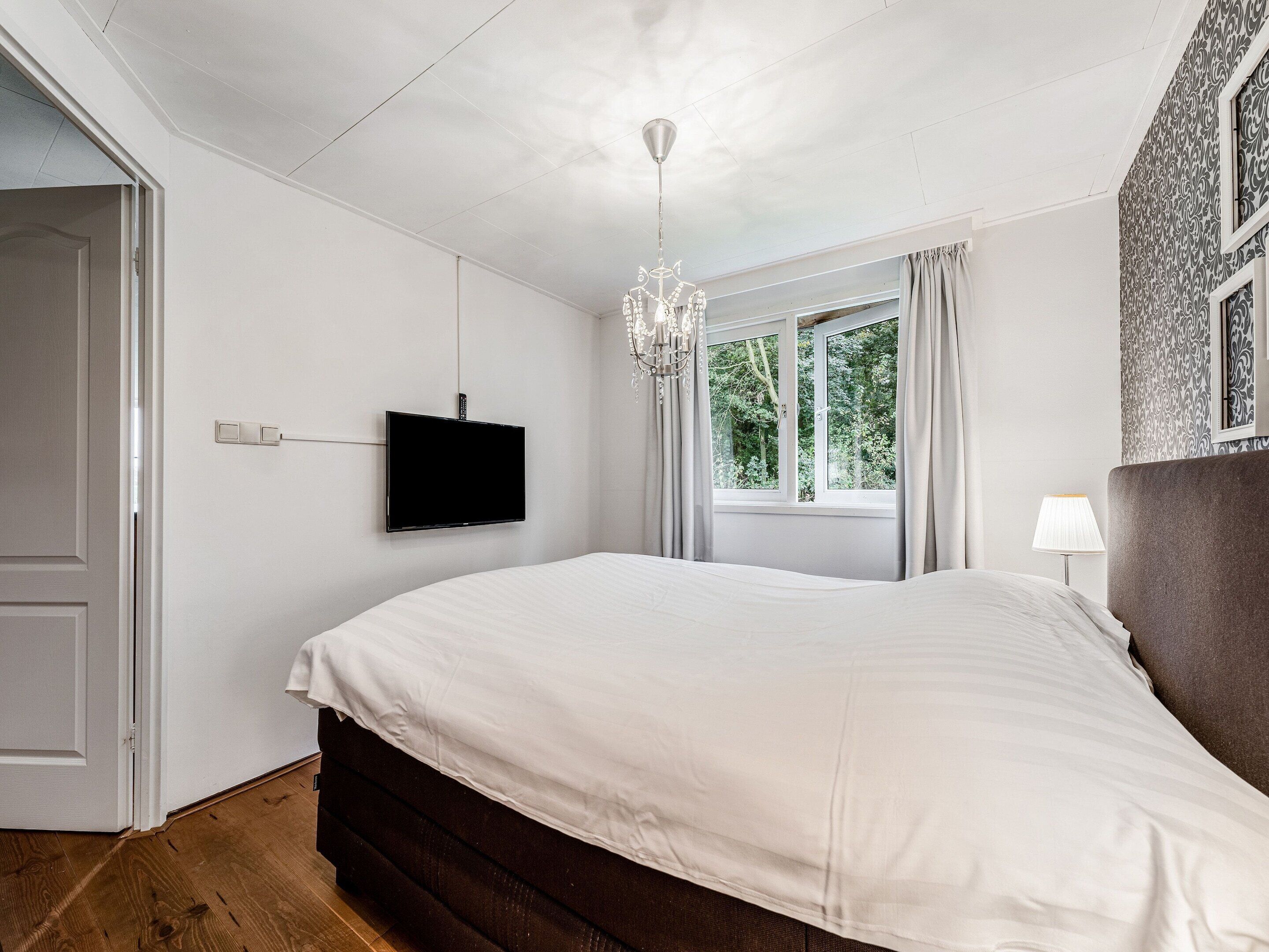 A bedroom with a bed, a TV mounted on the wall, a window with curtains, and a chandelier.