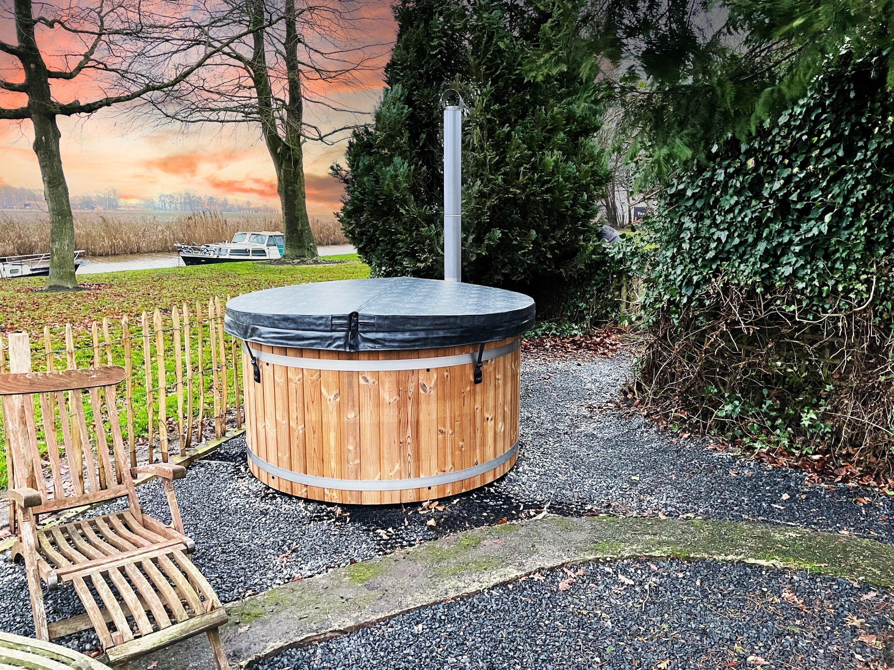 A wooden hot tub with a black cover, surrounded by a gravel path, a wooden chair, and a hedge.