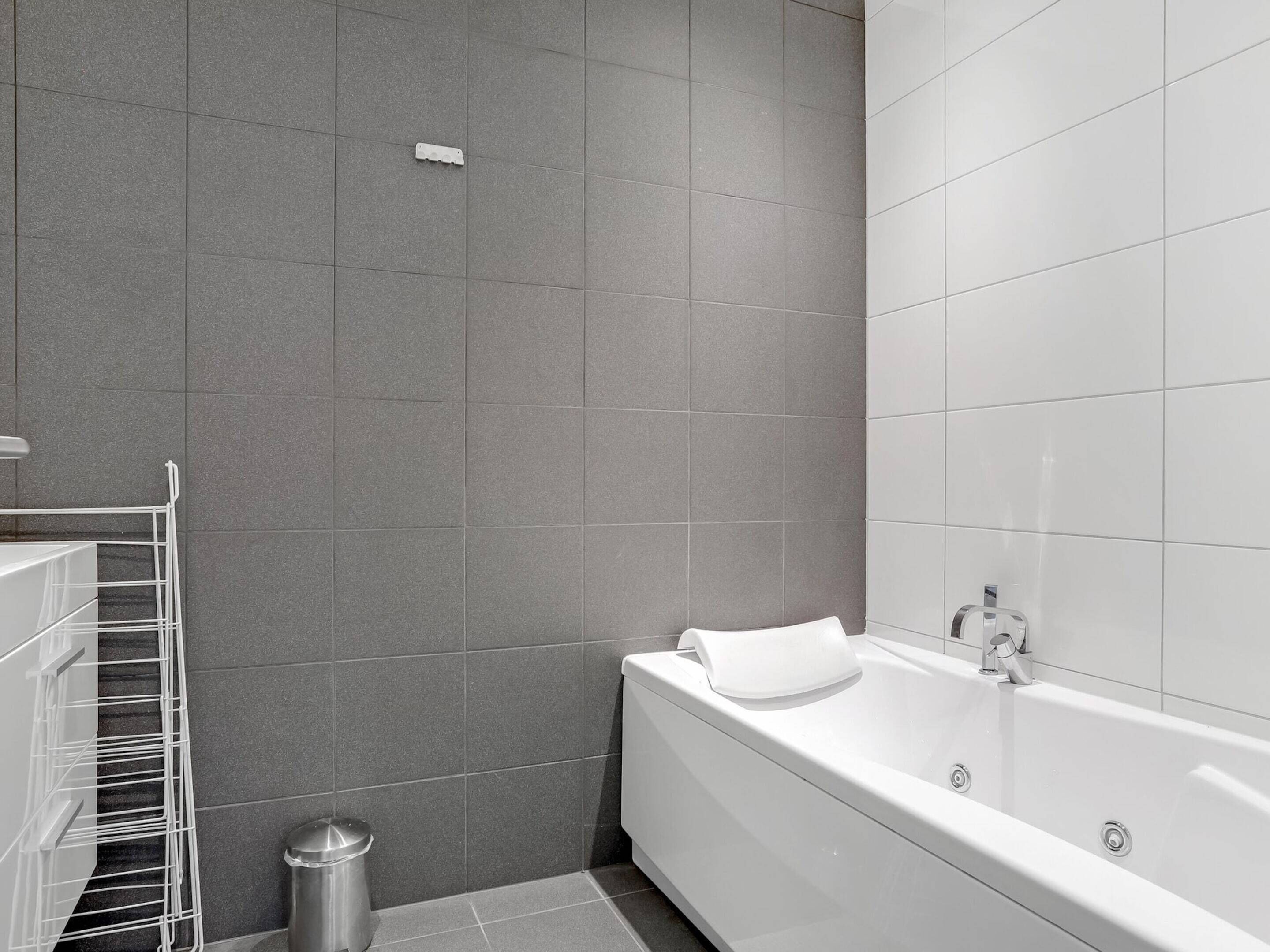 Un baño moderno con baldosas grises, una bañera blanca, una toalla blanca y un cubo de basura metálico.