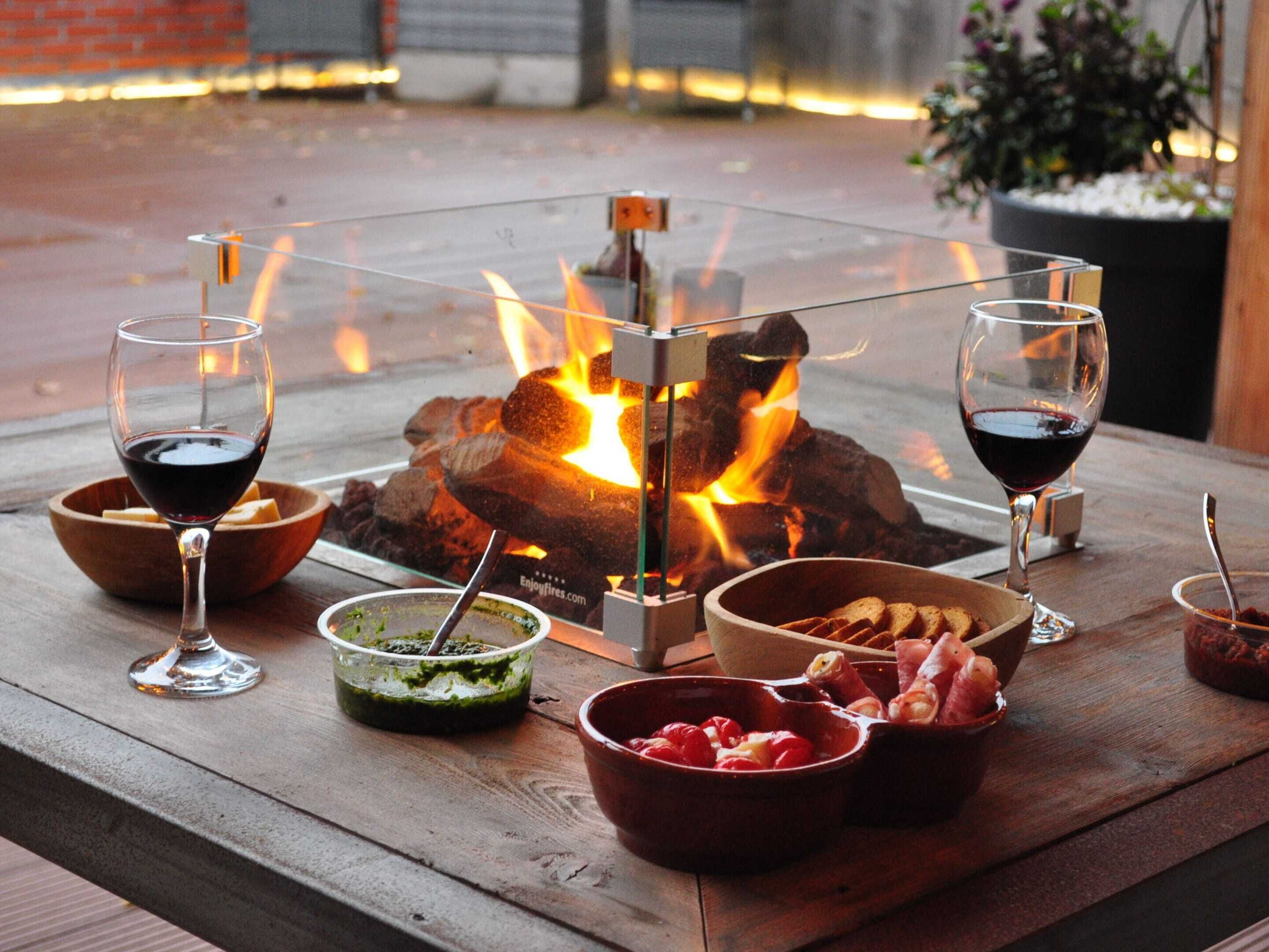 A table set outdoors with a fire feature, two glasses of red wine, and various food items.
